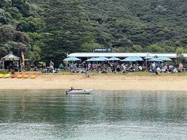 A lively beach scene at Otehei Bay with people enjoying various activities, including kayaking and dining under umbrellas.