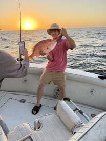 A young angler proudly displaying a large Red Snapper against a stunning sunset backdrop while fishing on a boat.