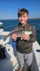 Young angler proudly holding a Speckled Trout while fishing on a boat in clear waters.