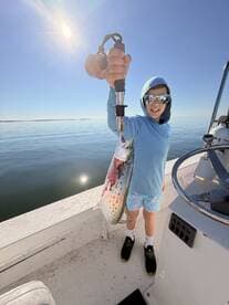 A young angler proudly displaying a freshly caught Speckled Trout while standing on a boat in calm waters.