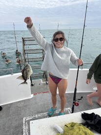 A young woman proudly displays her catch, a fish, while fishing on a boat, with pelicans visible in the background.