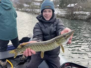 A young angler proudly displaying a Northern Pike while fishing from a boat in a scenic river setting.