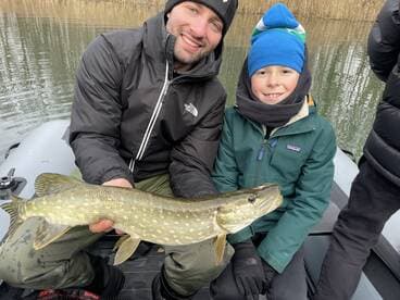 A young angler and an adult proudly display a Northern Pike while fishing from an inflatable boat in a serene waterway.