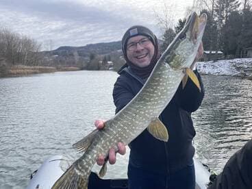 Angler proudly displaying a large Northern Pike while fishing on a scenic river during winter.