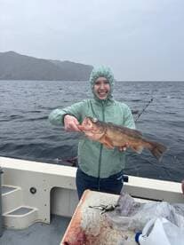 A young angler proudly displaying a large rockfish while fishing on a boat in a scenic coastal environment.