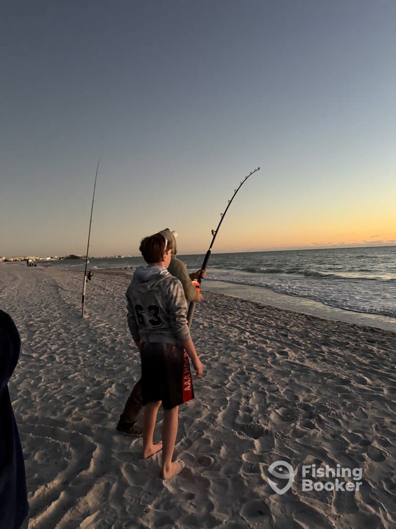 Two young anglers fishing on the beach at sunset, with one actively casting a line into the ocean.