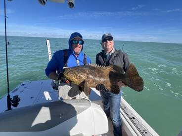Two anglers proudly displaying a large Goliath Grouper while fishing on a boat in clear waters.