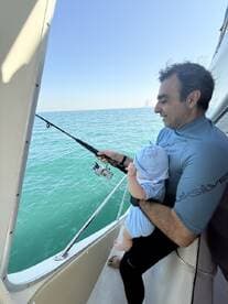 A father enjoys a fishing experience on a boat while holding his young child, with calm turquoise waters in the background.