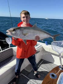 A young angler proudly displaying a large Amberjack while fishing on a boat in clear blue waters.