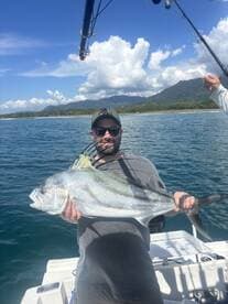 Angler proudly displaying a large Roosterfish while fishing off the coast, with a scenic backdrop of mountains and clouds.