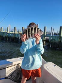 A young angler proudly displaying a striped Sheepshead fish while fishing from a boat near a dock.