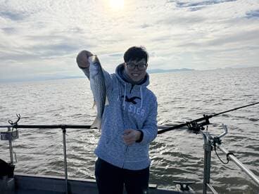 A young angler proudly displaying a striped bass while fishing on a boat, with a scenic water backdrop.