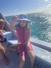 A young angler in a pink wetsuit proudly displaying a colorful fish while fishing on a boat in sunny waters.