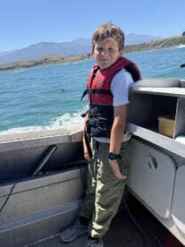 A young boy in a life jacket poses inside a fishing boat, enjoying a day on the water with scenic mountains in the background.