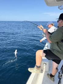 Two anglers actively fishing on a boat, one reeling in a fish while the other assists, showcasing a dynamic fishing experience in open waters.