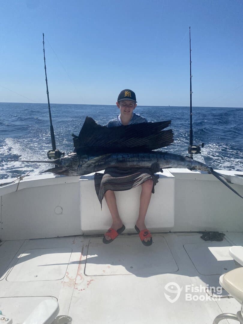 A young angler proudly displaying a large sailfish while sitting on the boat during a deep-sea fishing trip.