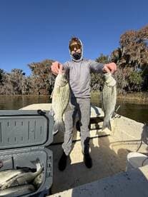 Young angler proudly displaying two large striped bass while fishing from a boat in a scenic river setting.
