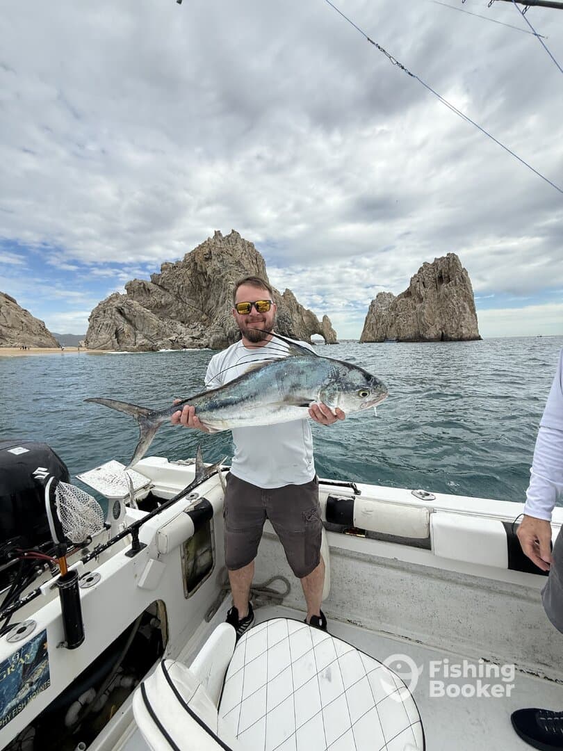 Angler proudly displaying a large fish, likely a Jack Crevalle, aboard a fishing boat near iconic rock formations.