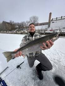 Angler proudly displaying a large Rainbow Trout while ice fishing on a snowy lake.