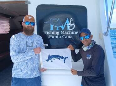 Two anglers proudly displaying a flag featuring a Marlin while standing in front of the Fishing Machine charter boat in Punta Cana.
