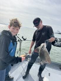 A young angler and an adult are on a boat, showcasing a caught stingray during a fishing trip.