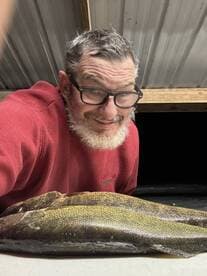 An angler proudly poses with two freshly caught Walleye on a table, showcasing a successful fishing trip.