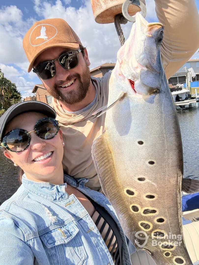 Two anglers proudly displaying a large Speckled Trout after a successful fishing trip, with a scenic waterfront backdrop.