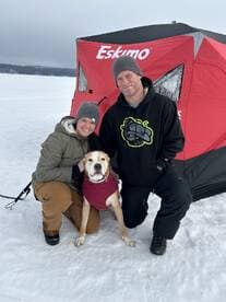 A couple poses with their dog in front of an ice fishing shelter on a snowy landscape.
