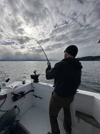 A fisherman actively reeling in a catch on a boat, surrounded by a scenic ocean backdrop under cloudy skies.