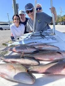 A cheerful group poses with their catch of the day, featuring a variety of fish including Snapper and Mackerel, displayed on a board after a successful fishing trip.