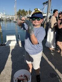 A young angler proudly displaying a caught fish at the marina, with a bucket of additional catches nearby.