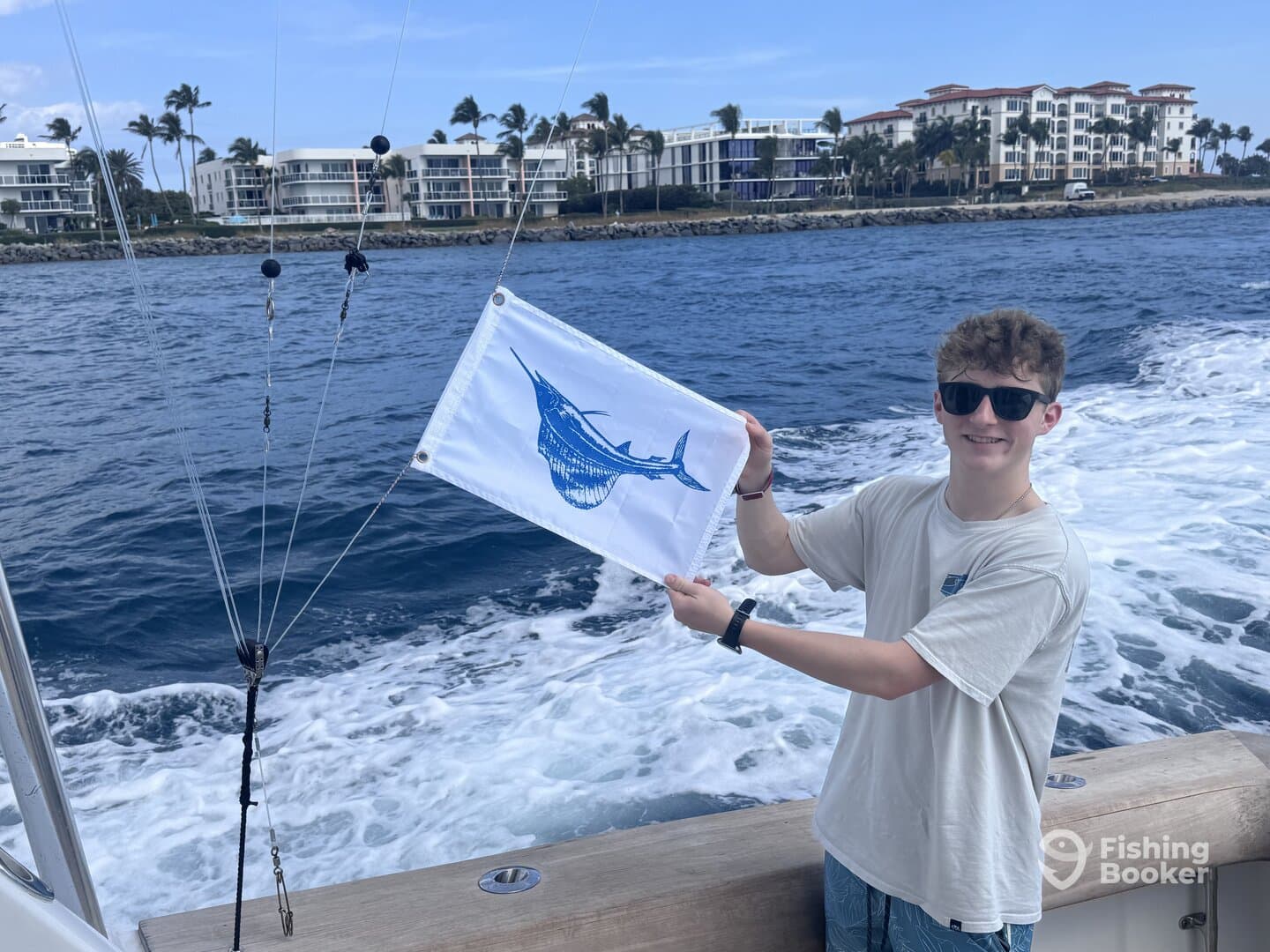 A young angler proudly displaying a flag featuring a marlin while on a fishing boat, with a scenic waterfront in the background.