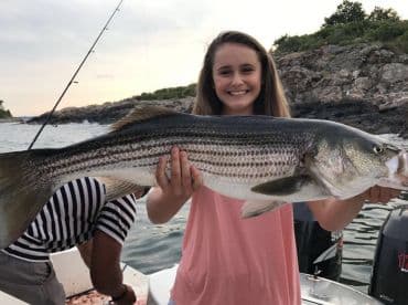 A young angler proudly displaying a large Striped Bass while fishing from a boat in a scenic coastal environment.