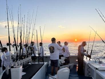 A group of anglers fishing at sunset on a charter boat, with multiple rods in action against a beautiful ocean backdrop.