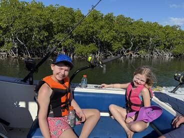 Two young anglers enjoying a day on a fishing boat, equipped with life jackets, surrounded by mangroves.