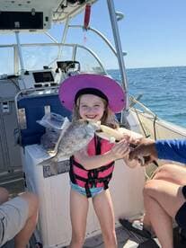 A young girl in a pink life jacket proudly holds a large fish while on a boat, showcasing her successful catch during a family fishing trip.