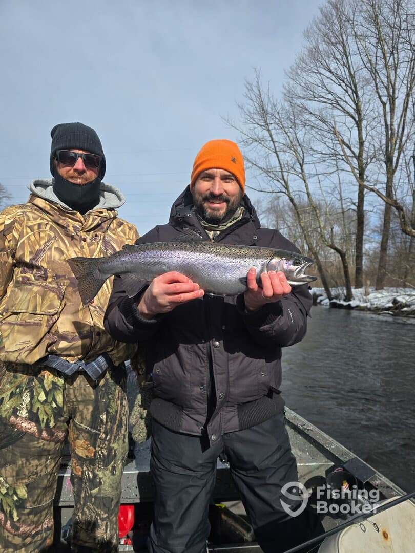 Two anglers proudly displaying a large Rainbow Trout while fishing in a river during winter.