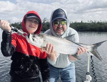 A young angler and an adult proudly displaying a large Tarpon while fishing in a scenic coastal environment.