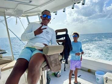 Two young anglers enjoying a day on a fishing boat, with one sitting in the helm chair and the other preparing fishing gear.