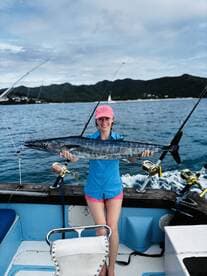 A young woman proudly displaying a large Wahoo caught while fishing on a boat, surrounded by a scenic ocean backdrop.
