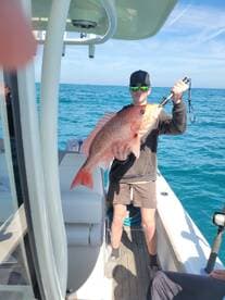 A young angler proudly displaying a large Snapper while fishing on a boat in clear blue waters.