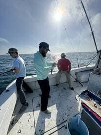 A group of anglers actively fishing on a boat, with one person reeling in a catch while others observe the action on a sunny day at sea.