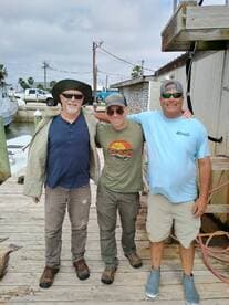 Three anglers posing together on a dock, with a fishing boat visible in the background.