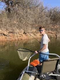 A young angler is actively fishing from a small boat, using a net to catch a fish in a serene river setting.