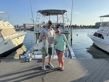 Two anglers proudly displaying their catch of two large fish at the dock, with a fishing boat in the background.