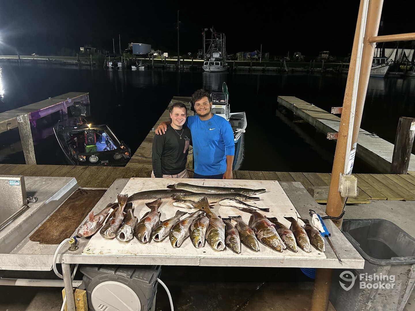 Two anglers proudly posing with a variety of fish, including Speckled Trout and Redfish, displayed on a cleaning table at a dock.