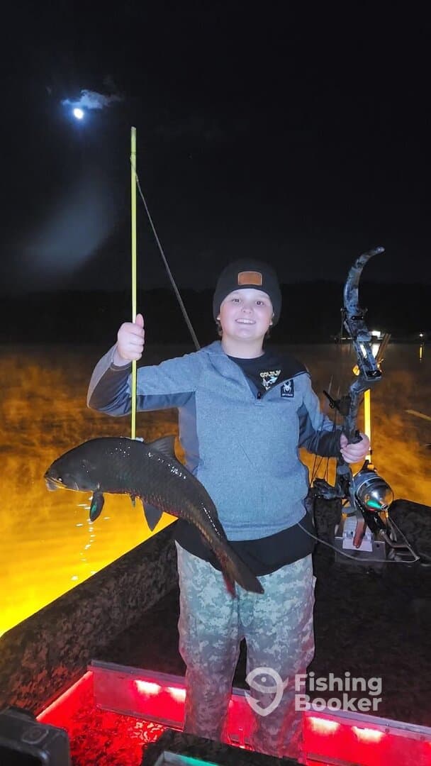 A young angler proudly displaying a caught fish while fishing at night, illuminated by the boat's lights.
