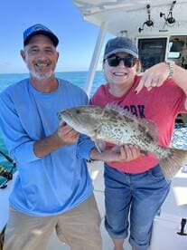 Two anglers proudly displaying a large Grouper caught during a fishing trip, with a vibrant ocean backdrop.