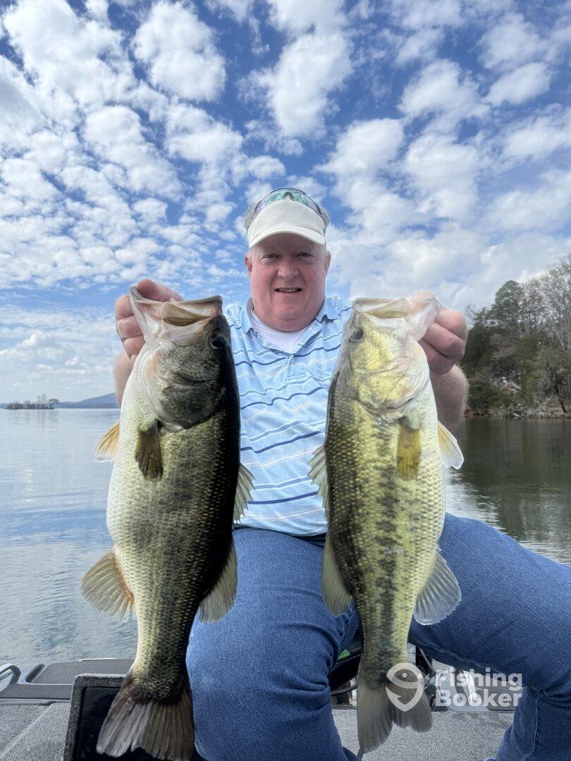 Angler proudly displaying two large Largemouth Bass while fishing on a serene lake under a beautiful sky.