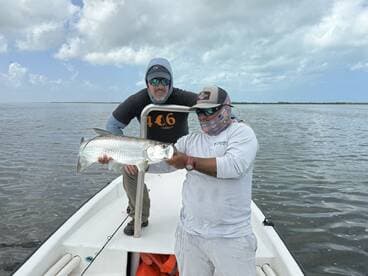 Two anglers proudly displaying a large Tarpon while fishing on a boat in a scenic coastal environment.